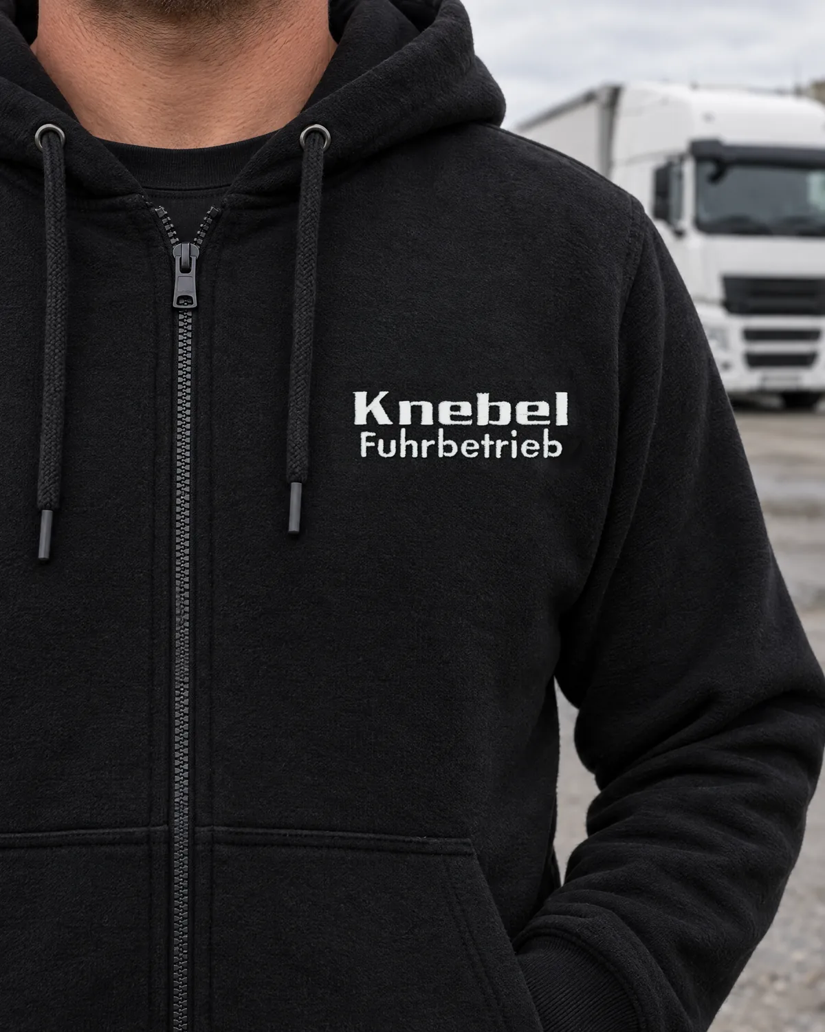 Fahrer im schwarzen Zip-Hoodie mit weiß gesticktem Schriftzug Knebel Fuhrbetrieb, Lkw im Hintergrund